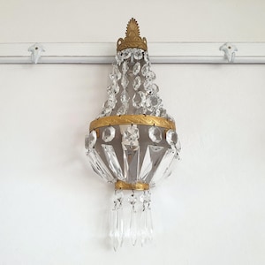 Puede incluir: Una l&aacute;mpara de ara&ntilde;a de cristal antigua con un marco de metal dorado. La l&aacute;mpara est&aacute; adornada con numerosos cristales transparentes y cuelga de una pared blanca. El dise&ntilde;o es ornamentado y elegante, adecuado para la decoraci&oacute;n del hogar.