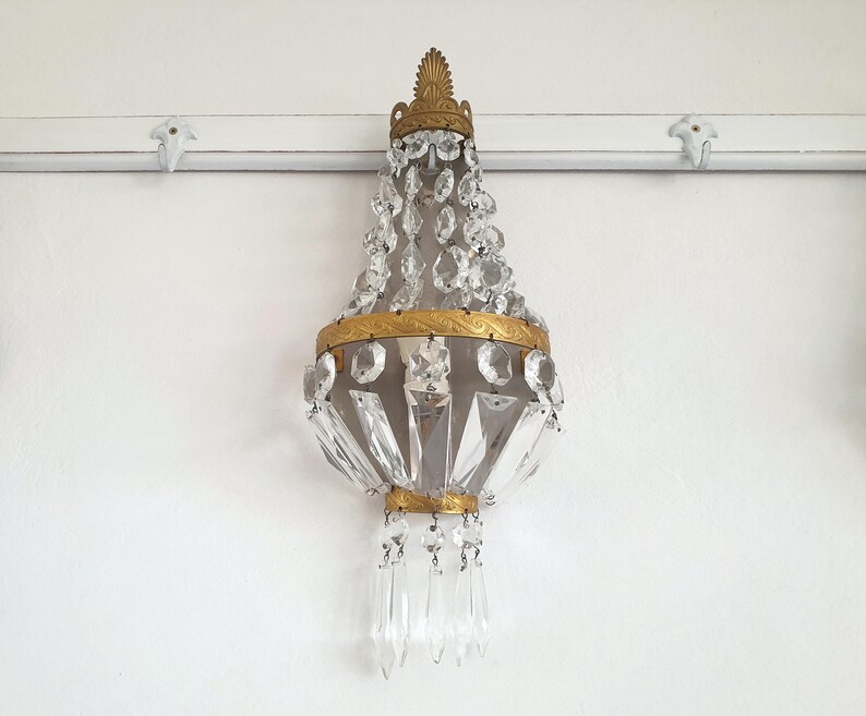 Puede incluir: Una l&aacute;mpara de ara&ntilde;a de cristal antigua con un marco de metal dorado. La l&aacute;mpara est&aacute; adornada con numerosos cristales transparentes y cuelga de una pared blanca. El dise&ntilde;o es ornamentado y elegante, adecuado para la decoraci&oacute;n del hogar.