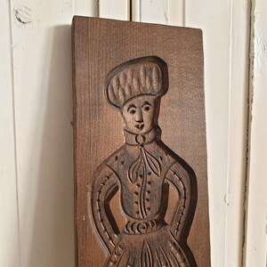 Puede incluir: Un molde de galletas de madera rectangular vintage con una imagen tallada de una persona con vestimenta tradicional. La figura lleva un sombrero, una camisa abotonada y una falda larga. El molde es de color marr&oacute;n oscuro.