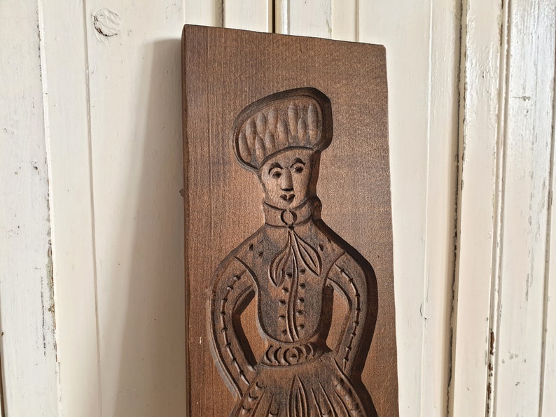 Puede incluir: Un molde de galletas de madera rectangular vintage con una imagen tallada de una persona con vestimenta tradicional. La figura lleva un sombrero, una camisa abotonada y una falda larga. El molde es de color marr&oacute;n oscuro.
