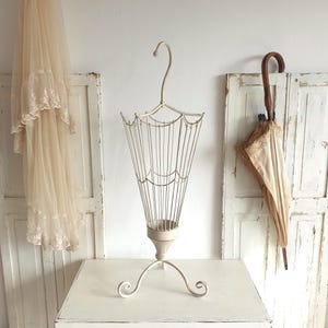 May include: An off-white metal umbrella stand with a decorative, cage-like design. The stand has a curved handle and sits on a three-legged base. A vintage umbrella with a wooden handle and lace trim is leaning against a white door.