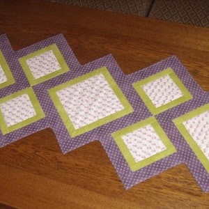 Op de afbeelding: Een paarse en groene tafelrunner met een ruitpatroon. De runner is gemaakt van stof met een bloemenprint en heeft een gestikte textuur.