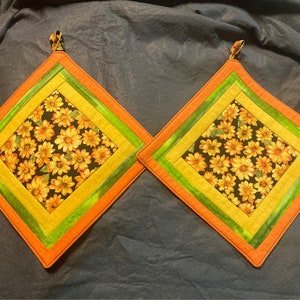 Könnte beinhalten: Zwei gequiltete Topflappen mit einem Sonnenblumenmuster in der Mitte. Die Topflappen sind orange, gelb und grün mit schwarzem Hintergrund.
