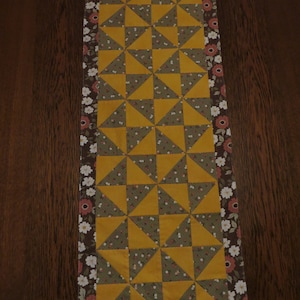 May include: A table runner with a geometric pattern of yellow and green triangles. The runner has a brown and white floral border.