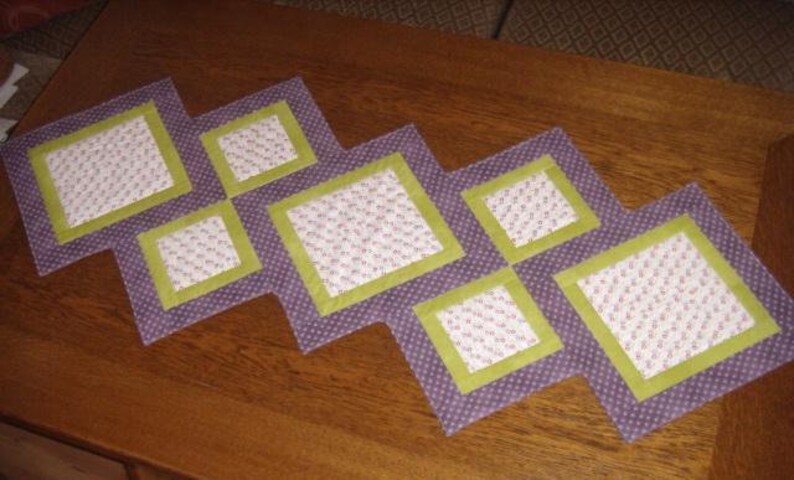 Op de afbeelding: Een paarse en groene tafelrunner met een ruitpatroon. De runner is gemaakt van stof met een bloemenprint en heeft een gestikte textuur.