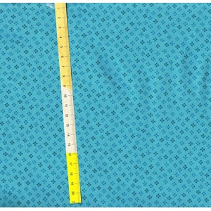 Könnte beinhalten: Türkisfarbener Stoff mit sich wiederholendem Blumenmuster. Ein gelbes Lineal ist vertikal auf dem Stoff platziert und zeigt Messungen bis zu 50 cm an. Der Stoff hat eine subtile Textur und eine gleichmäßige Farbe.