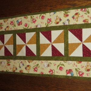 Peut inclure: Un chemin de table avec un motif floral dans des tons de jaune, vert et marron. Le chemin de table présente un panneau central de six carrés alternés avec un motif de moulin à vent en blanc, bordeaux et or.