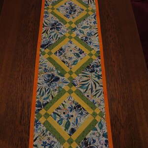 Könnte beinhalten: Ein Tischläufer mit einem floralen Muster in Blautönen, Grün und Gelb. Der Läufer hat ein Diamantenmuster mit einem gelben und grünen Rand. Der Läufer liegt auf einem Holztisch.