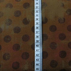 Op de afbeelding: Bruine stof met een stippenpatroon. De stippen zijn donkerbruin van kleur en hebben een verweerde uitstraling. Een witte meetlint is zichtbaar aan de rechterkant van de afbeelding. De lint meet van 29 tot 45 inch.