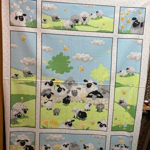 May include: Fabric panel featuring a whimsical sheep design. The fabric showcases cartoon sheep in a green pasture with blue skies, clouds, and bees. The sheep are white and black, with some gray sheep. The panel is divided into rectangular sections.