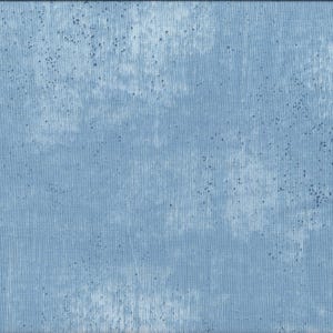 Op de afbeelding: Een lichtblauwe stof met een getextureerde, verweerde uitstraling. De stof heeft een gevlekt effect met donkerblauwe spikkels en verticale strepen. Een gele liniaal met zwarte markeringen is langs de rechterrand geplaatst, tot 50 cm.