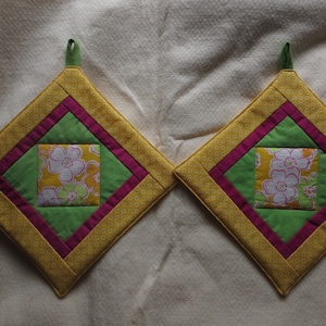 Könnte beinhalten: Zwei gelbe, grüne und rosafarbene gequiltete Topflappen mit floralem Stoff in der Mitte. Die Topflappen haben eine Diamantform und eine Schlaufe zum Aufhängen.