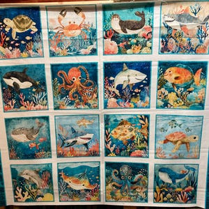Könnte beinhalten: Ein Stoffpaneel mit farbenfrohen Aquarellillustrationen verschiedener Meeresbewohner, darunter eine Schildkröte, eine Krabbe, ein Hai und ein Oktopus. Das Kunstwerk ist vor einem blauen Hintergrund mit Korallen- und Seetangdetails angeordnet und erzeugt eine Unterwasserszene. Das Panel hat einen weißen Rand mit einem Wellenmuster.