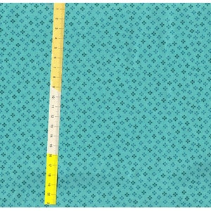 Op de afbeelding: Teal stof met een herhalend patroon van kleine, donker teal klavertjes met vier blaadjes. Een gele liniaal met zwarte markeringen is verticaal geplaatst, met metingen in centimeters.
