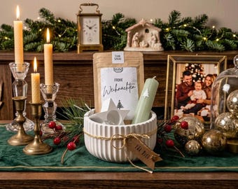 May include: A white ribbed ceramic bowl containing a brown paper pouch labeled Frohe Weihnachten, a sage green candle with a white tree icon, and a white star-shaped candle holder, tied with twine and a tag reading Für Dich.