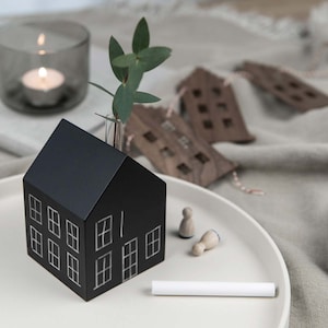 Puede incluir: Un jarrón negro con forma de casa con contornos de ventanas blancos, que contiene ramitas de eucalipto en un pequeño tubo de vidrio. Dos figuras de madera y un trozo de tiza blanca están sobre un plato blanco. Una vela encendida en un portavelas de vidrio está en el fondo.