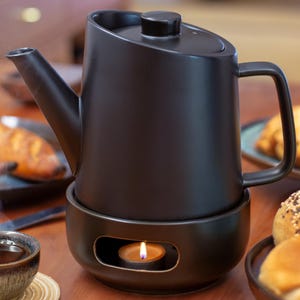 Peut inclure: Une théière en céramique noire avec un chauffe-eau intégré est posée sur une table en bois avec une bougie allumée à l'intérieur. La théière a une poignée et un bec verseur. Il y a des pâtisseries sur la table à côté de la théière.