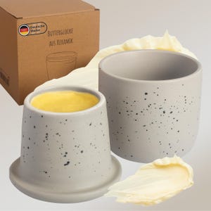 Puede incluir: Una mantequera de cerámica gris con motas negras, llena de mantequilla amarilla. Junto a ella, un recipiente vacío a juego y una mancha de mantequilla. Una caja de cartón con texto alemán en el fondo.