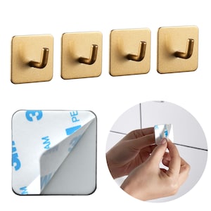 Könnte beinhalten: Vier goldfarbene, quadratische Wandhaken mit gebogenem Haken-Design. Das Bild zeigt auch ein quadratisches Klebepad, bei dem die Rückseite teilweise abgezogen ist, und eine Person, die die Rückseite von einem kleineren Klebepad abzieht.