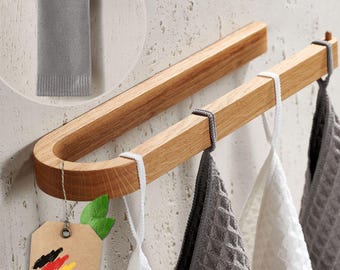 May include: A wooden towel rack with a curved design and four hooks. The rack is mounted on a wall and has three towels hanging from it. The towels are white and gray. The rack is made of natural wood and has a rustic look. The text "Deutsche Marke" is visible on a brown tag.
