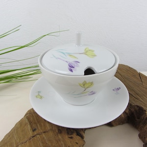 Op de afbeelding: Een witte keramische suikerpot met deksel en schotel, versierd met paarse en gele bloemmotieven. De kom heeft een klein handvat op het deksel en een sleuf voor de lepel. De set staat op een houten oppervlak.