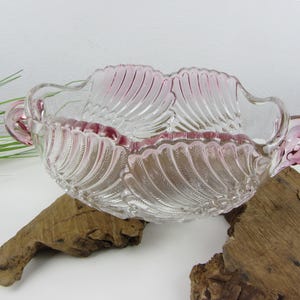 Könnte beinhalten: Eine klare Glasschüssel mit rosa Akzenten und einem gewellten Rand. Die Schüssel hat zwei Griffe und ein federartiges Design.