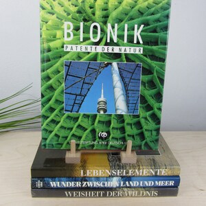 Op de afbeelding: Een stapel van drie hardcover boeken met groene, blauwe en bruine omslagen. Het bovenste boek heeft de titel "BIONIK PATENTE DER NATUR" en een afbeelding van een toren met een groen bladpatroon. Het tweede boek heeft de titel "LEBENSELEMENTE" en het derde boek heeft de titel "WUNDER ZWISCHEN LAND UND MEER". Het onderste boek heeft de titel "WEISHEIT DER WILDNIS".