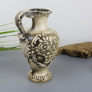 May include: A small, cream-coloured ceramic vase with a brown floral design. The vase has a handle and a round base.