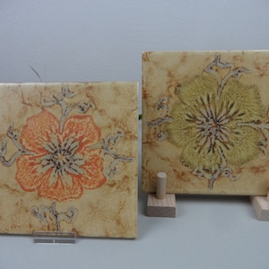 May include: Two ceramic tiles featuring floral designs. The tile on the left has a large orange flower with a brown centre and a beige background. The tile on the right has a large yellow flower with a brown centre and a beige background.