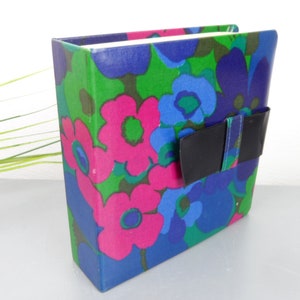 Könnte beinhalten: Ein Vintage-Fotoalbum mit einem farbenfrohen Blumenmuster in Pink, Blau und Grün. Das Album hat einen schwarzen Verschluss. Das Cover ist eine lebendige Mischung aus Retro-Farben und -Designs. Das Innere des Albums ist weiß.