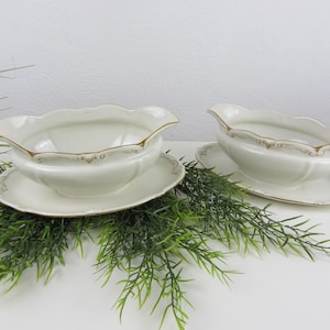 Könnte beinhalten: Zwei weiße Porzellan-Saucieren mit goldenen Verzierungen und passenden Untertellern. Die Saucieren haben einen gewellten Rand und ein zartes Blumenmuster.