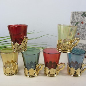 Könnte beinhalten: Ein Set aus sechs kleinen Gläsern in verschiedenen Farben wie Rot, Gelb, Grün und Blau. Jedes Glas befindet sich in einem dekorativen goldfarbenen Metallhalter mit einem detaillierten Blattmuster und einem kleinen Henkel an der Basis.