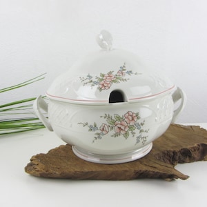 Peut inclure: Une soupière en céramique blanche avec couvercle assorti, ornée d'un motif floral de roses roses et de petites fleurs bleues. Elle possède deux anses latérales, une encoche pour louche et un fin liseré rose sur la base et le couvercle.