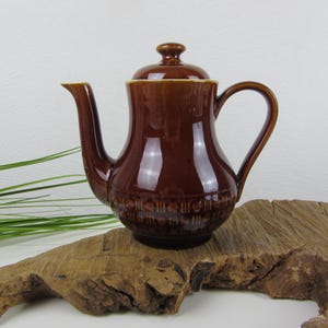 Op de afbeelding: Een bruine keramische theepot met een deksel en een handvat. De theepot heeft een glanzende afwerking en een decoratieve band rond de basis. Het wordt gepresenteerd op een stuk drijfhout.