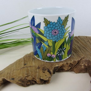 Könnte beinhalten: Ein weißer Keramikübertopf mit einem lebendigen botanischen Druck aus großen blauen Blumen, grünem Blattwerk und kleinen rosa Akzenten. Das zylindrische Gefäß hat eine glatte Oberfläche und eine saubere, offene Oberseite mit Grafik.