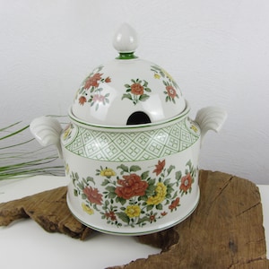 Op de afbeelding: Een witte porseleinen soepterrine met een deksel, met een bloemenpatroon in tinten rood, geel en groen. De terrine heeft twee handvatten en een decoratieve groene en witte rand.