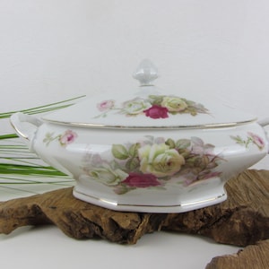 Pode incluir: Uma travessa de porcelana branca com tampa, adornada com padrões florais de rosas em tons de rosa, amarelo e verde. A travessa tem acabamentos e alças douradas e é exibida em madeira flutuante.