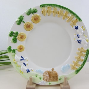 Könnte beinhalten: Weiße Keramikplatte mit einem Bauernhof-Szenen-Design. Der Rand zeigt Sonnenblumen, grüne Blätter, einen gelben Zaun und weiße Enten mit blauen Akzenten. Eine braune Scheune und blaue Wolken sind ebenfalls abgebildet.