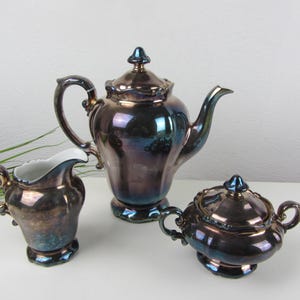 Op de afbeelding: Een vintage theeservies met een metallic afwerking, bestaande uit een theepot, een melkkan en een suikerpot. De set heeft een donkere, iriserende glans met blauwe en paarse tinten. De melkkan heeft een witte binnenkant.