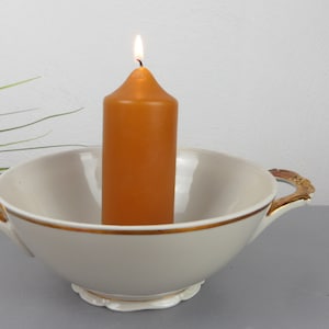 May include: A lit orange candle sits in a cream-coloured bowl with gold trim and handles. The bowl has a scalloped base. The candle is centred in the bowl and has a lit flame.