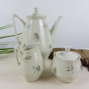 May include: A vintage porcelain tea set featuring a teapot, creamer, and sugar bowl. The set is off-white with delicate floral and leaf patterns in muted colors. The teapot has a tall, slender shape with a lid and a curved handle.