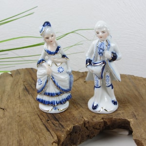 May include: Two porcelain figurines depicting a man and woman in 18th-century dress. The figures are white with blue and gold detailing. The woman wears a dress with a bow, and the man holds a drum.