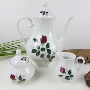 May include: A white porcelain tea set featuring a teapot, sugar bowl, and creamer. Each piece is decorated with a red rose and green leaves. The teapot has a lid and a curved handle. The sugar bowl has a lid and the creamer has a handle.