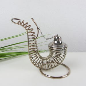 May include: A silver-toned metal tea infuser with a unique horn-like design. The infuser features a coiled body, a circular base, and a small, lidded compartment. A chain connects the lid to the coiled structure. The infuser is displayed on a white surface.