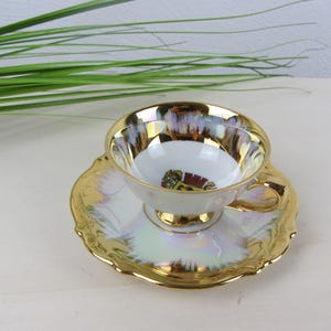 May include: A white teacup and saucer set with gold trim and iridescent detailing. The teacup has a small emblem inside. The saucer has a scalloped edge. The set is on a light-coloured surface.