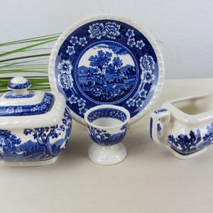 Könnte beinhalten: Eine Sammlung blau-weißer Keramik-Tafelservice, darunter ein Teller, eine Zuckerdose, ein Sahnekännchen und ein Eierbecher. Die Teile weisen ein Landschaftsdesign und florale Akzente auf, was eine klassische, Vintage-Ästhetik schafft.
