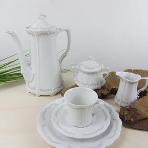 Op de afbeelding: Een wit porseleinen theeservies met een decoratief ontwerp. De set bevat een koffiepot, een suikerpot, een melkkan, een kopje, een schotel en een bord. De set wordt gepresenteerd op een licht oppervlak.