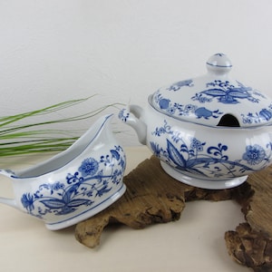 Può includere: Una salsiera e un piatto da portata in ceramica bianca con motivi floreali blu. La salsiera ha un manico e un beccuccio. Il piatto da portata ha manici e un pomello sul coperchio. Entrambi sono su un pezzo di legno.