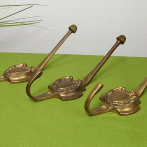 May include: Three antique brass coat hooks. Each hook has a decorative base with a curved hook and a long stem ending in a rounded knob. The hooks are arranged on a green surface, suggesting a vintage or retro theme.
