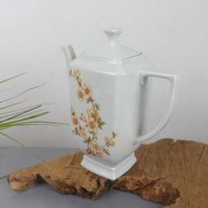 Peut inclure: Une théière en céramique blanche avec une base et un couvercle carrés, ornée d'un motif floral dans les tons de jaune et de marron. La théière a une anse et un bec. Elle est présentée sur un morceau de bois.